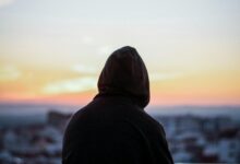 A solitary figure in a hoodie gazes at a vibrant sunset from a rooftop, creating a peaceful silhouette.