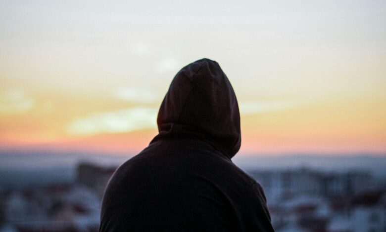 A solitary figure in a hoodie gazes at a vibrant sunset from a rooftop, creating a peaceful silhouette.