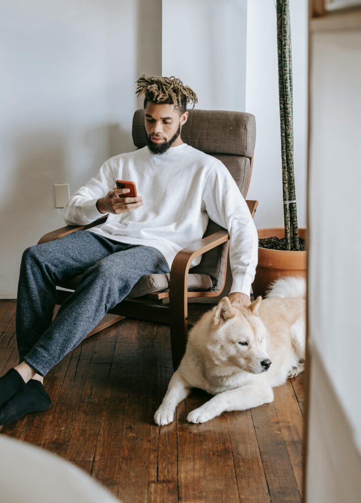 Man sitting in chair indoors texting on cellphone with a dog beside him